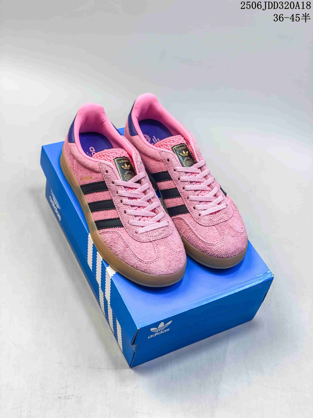 Pair of pink Adidas Gazelle sneakers displayed on a blue Adidas shoebox, showing black stripes, pink laces and gum-colored soles.