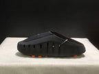 Side profile of a black slide showing vertical side openings, a textured upper, and orange outsole details.