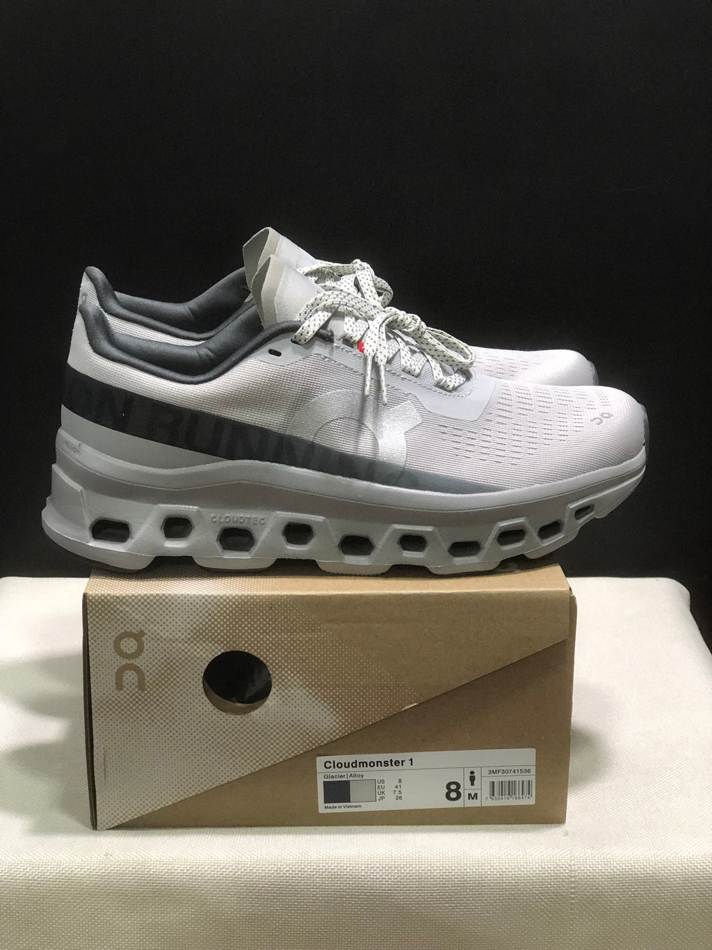 Side view of the On Cloudmonster 1 pair showing the dark heel band, lace-up upper, and cutout-style white outsole.