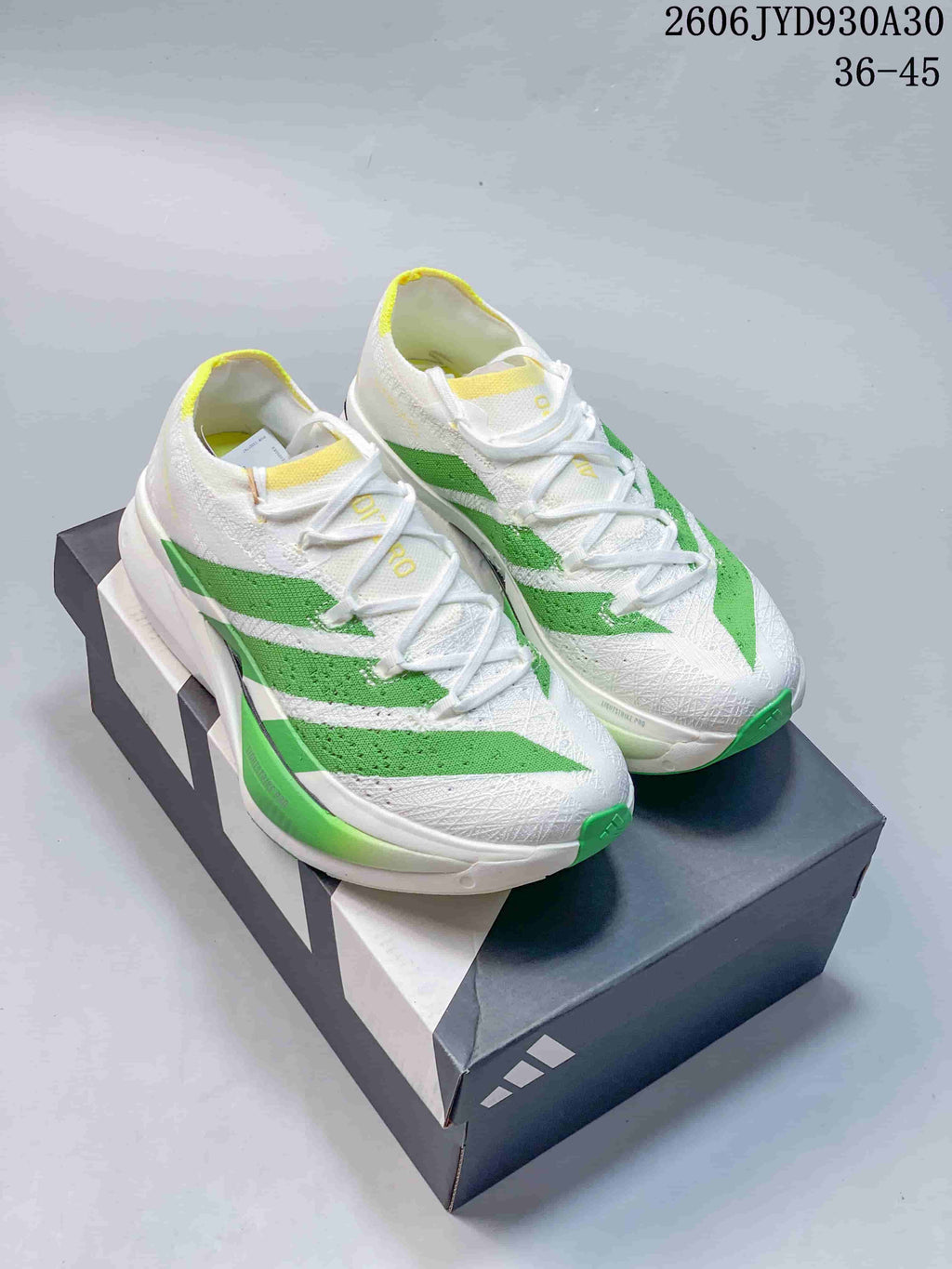 Pair of white adidas Adizero Prime X3 running shoes with green stripes and yellow accents, shown on a shoebox.