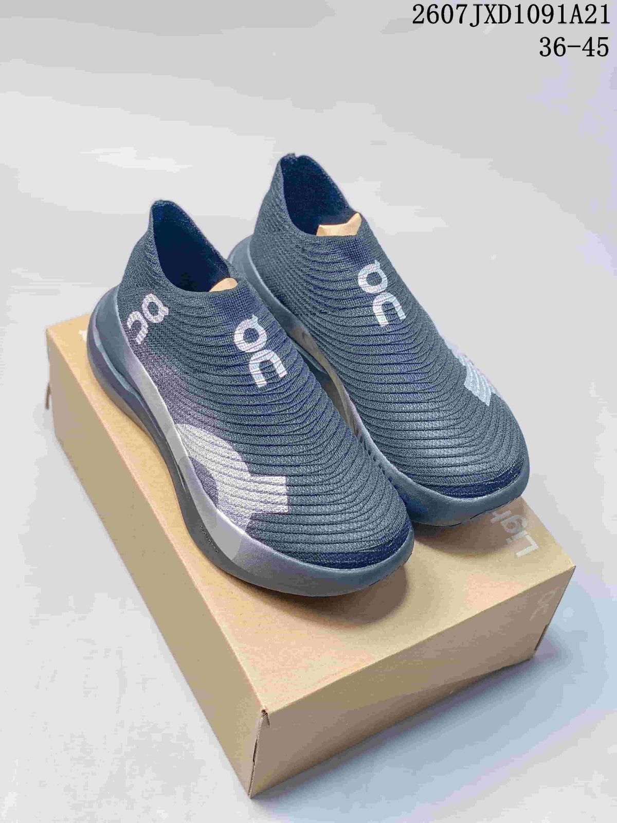 Pair of dark gray On Cloudboom Strike LS slip-on sneakers displayed on a shoebox, with white branding and side accents.