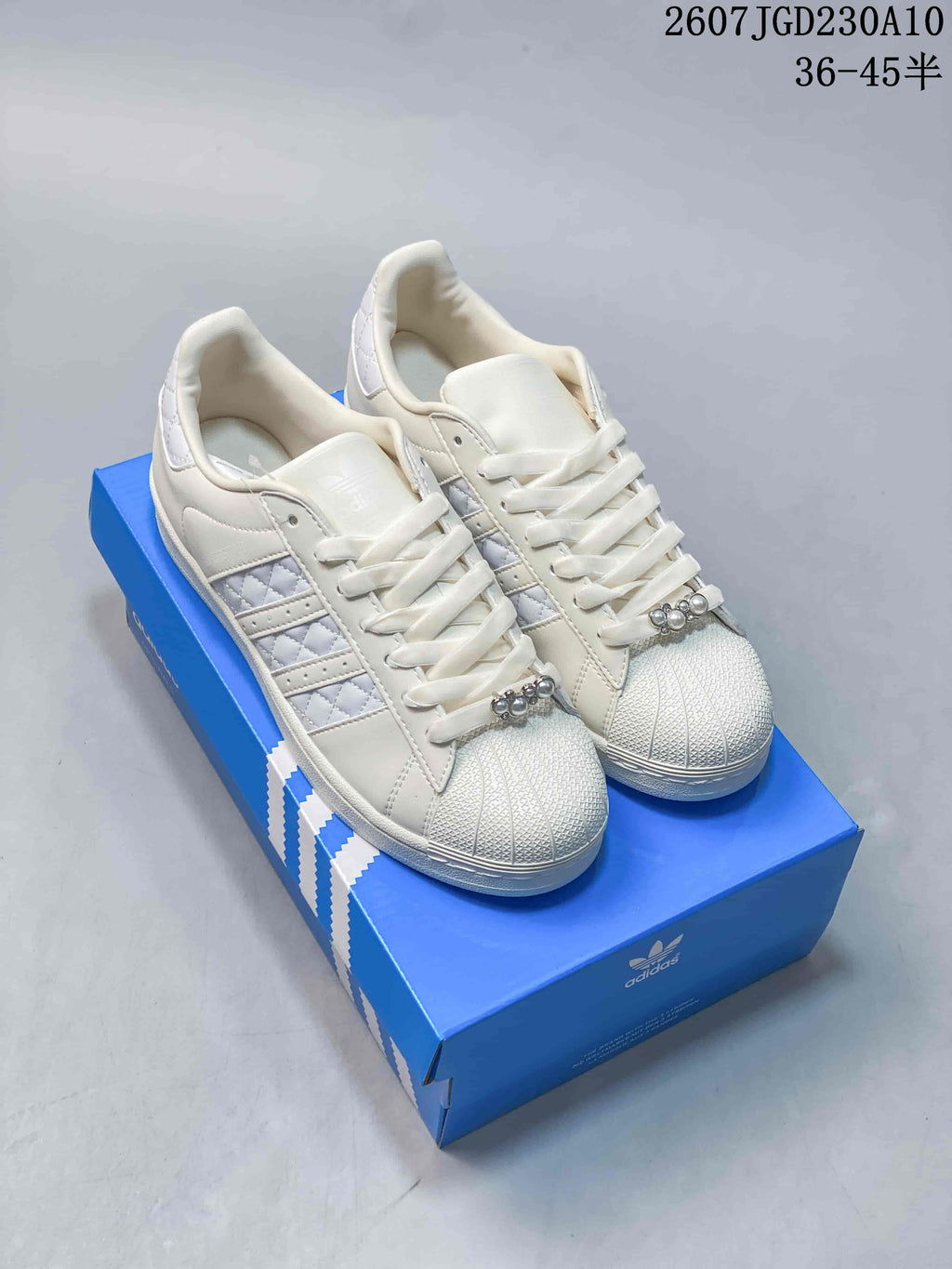 Pair of white Adidas Superstar II sneakers displayed on a blue shoebox, showing the shell toes, laces, side stripes, and metallic lace accents.