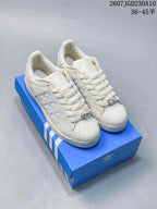 Pair of white Adidas Superstar II sneakers displayed on a blue shoebox, showing the shell toes, laces, side stripes, and metallic lace accents.