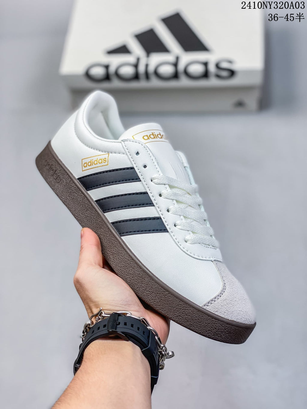 Angled side view of a white Adidas sneaker with black stripes, gray toe and heel panels, gold-toned branding, and a brown outsole.