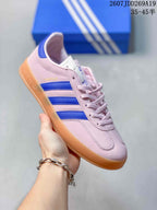 Side view of a pink Adidas Gazelle sneaker with blue stripes, lace-up closure, and gum-colored outsole.