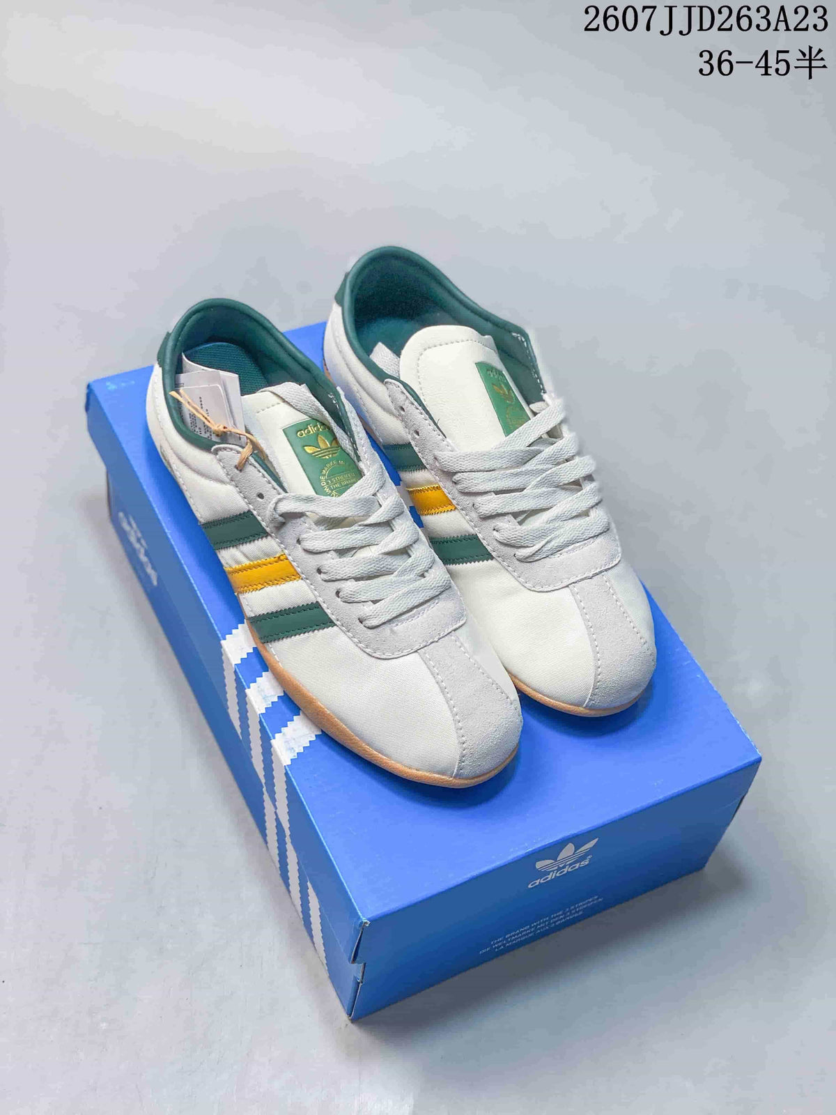 Pair of white adidas Originals Tokyo sneakers with green and yellow stripes displayed on a blue adidas shoe box.