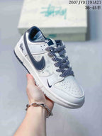 Angled side view of the white and dark Nike SB Dunk Low sneaker showing the Swoosh, patterned side panel, laces, and heel graphic.