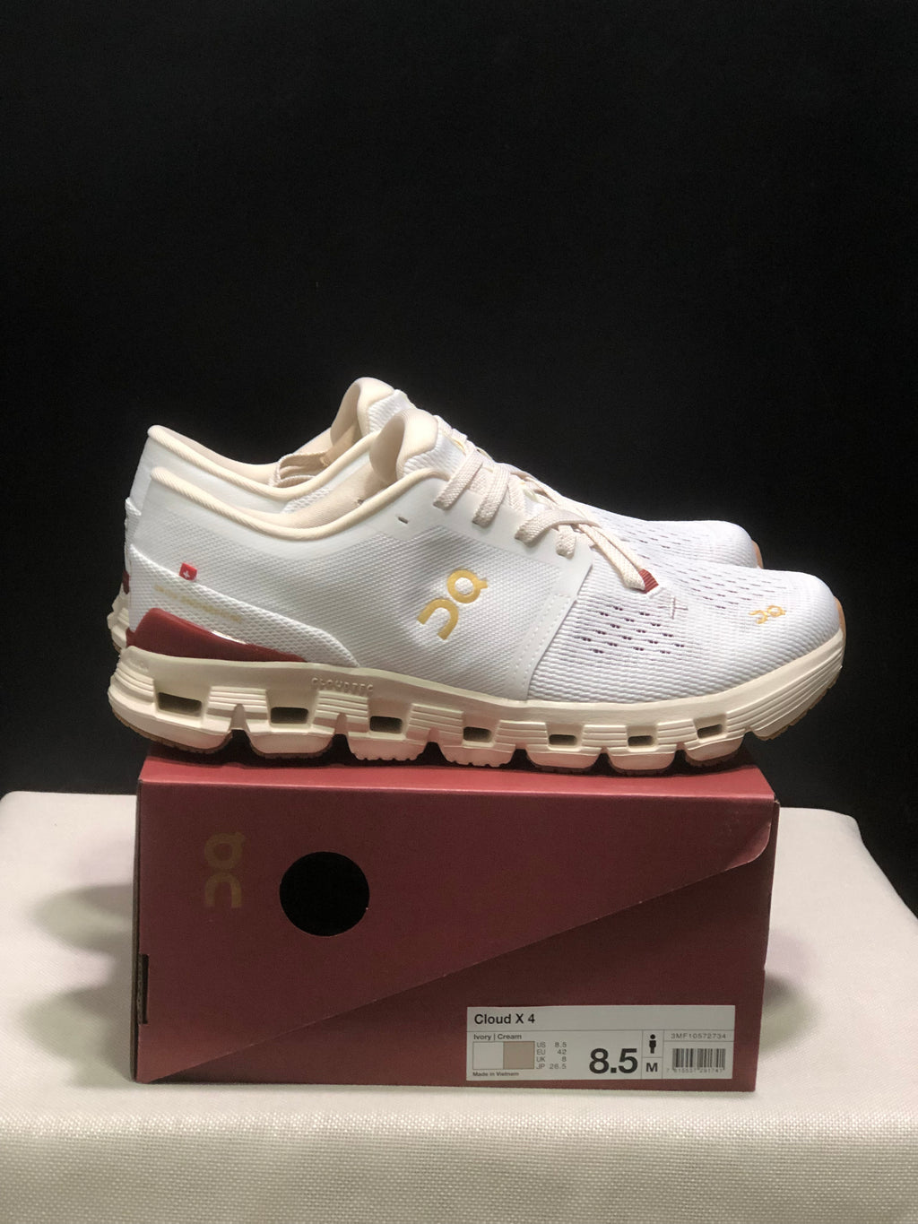 Side view of a white On Cloud X4 shoe with gold branding, burgundy heel detail, and a segmented cream outsole.