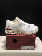 Side view of a white On Cloud X4 shoe with gold branding, burgundy heel detail, and a segmented cream outsole.