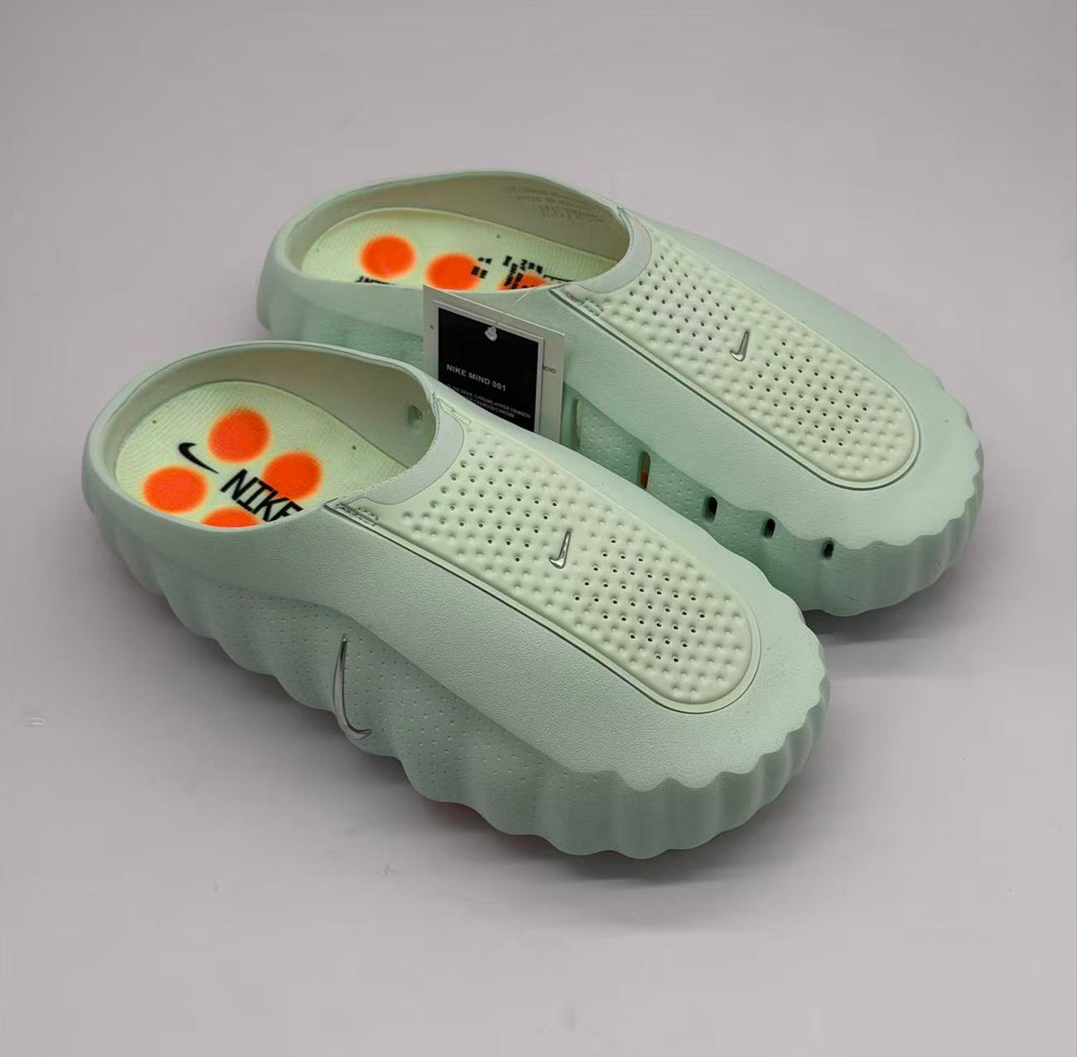 Top three-quarter view of two light green slides with perforated footbed panels, orange footbed graphics, and a visible product tag reading “NIKE MIND 001”.
