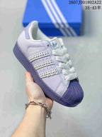 Close-up side view of a light lavender Adidas Superstar sneaker showing the SUPERSTAR text, trefoil tongue label, white laces, and decorative side stripes.