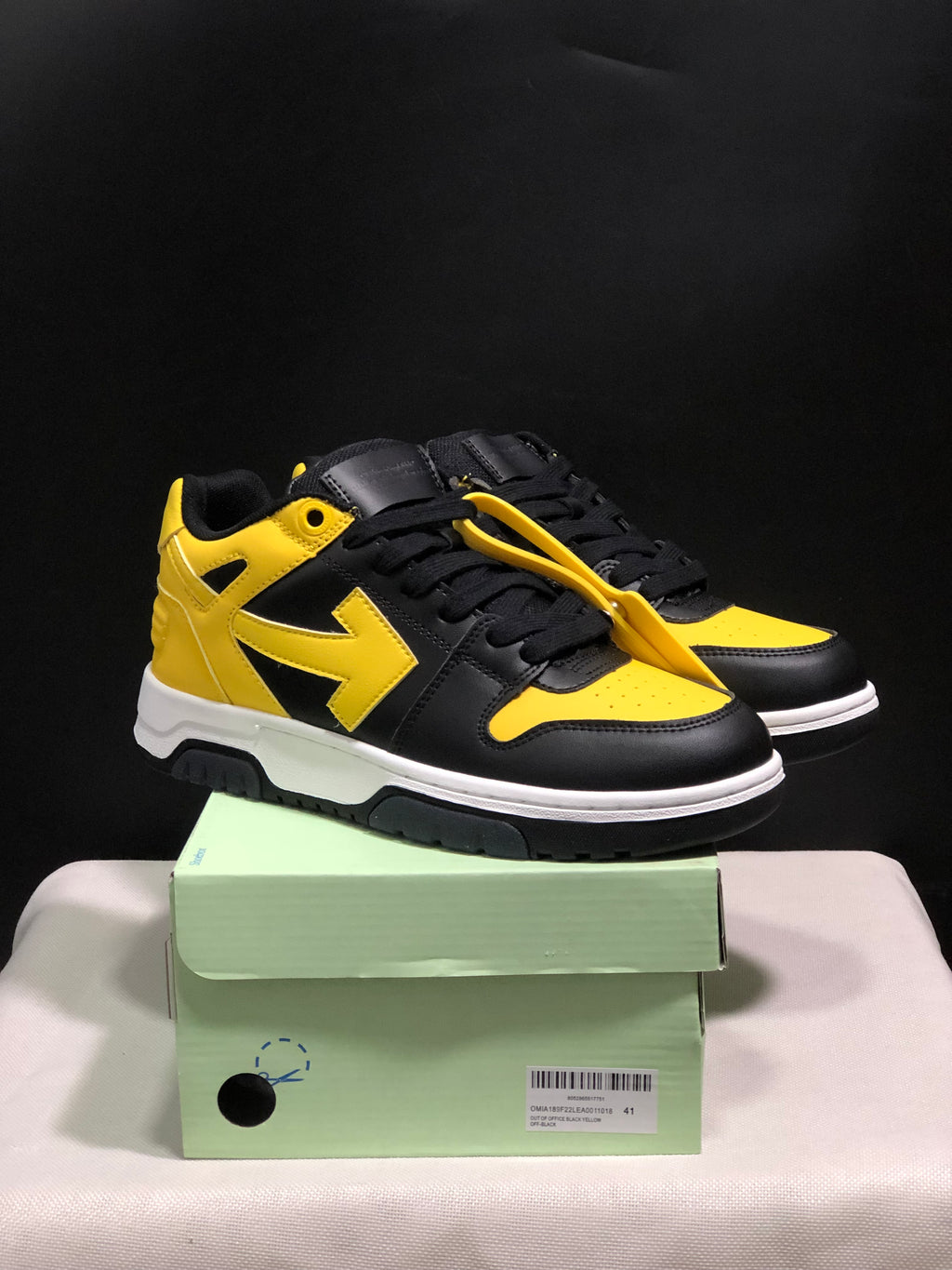 Pair of black and yellow low-top sneakers displayed on a green shoe box, showing the front and outer side.