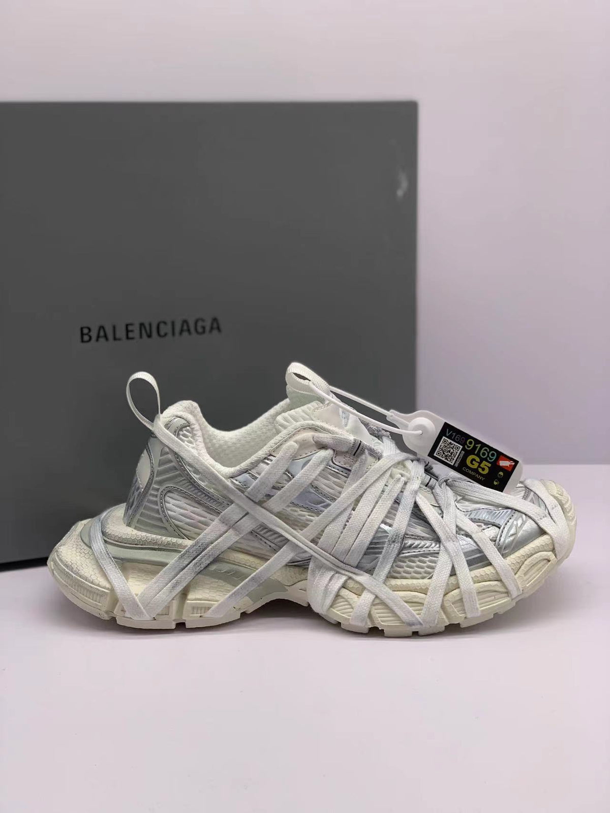Side view of all-white Balenciaga 3XL sneakers with layered laces and a branded box in the background.