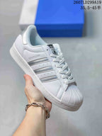 White adidas Superstar sneaker with silver serrated stripes and a textured shell toe; style code and size range are overlaid at the top.