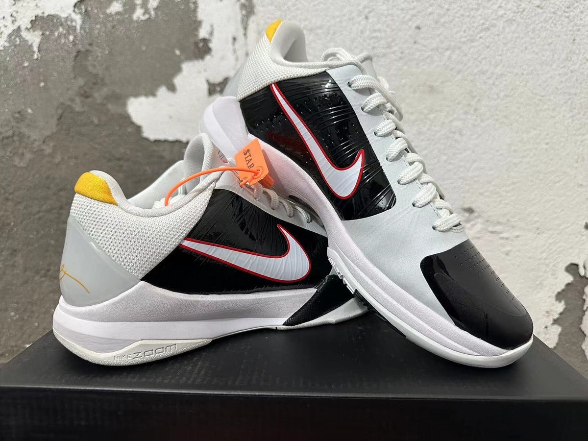 Two white and black Nike Kobe basketball shoes displayed together, showing the side Swoosh and yellow heel accents.