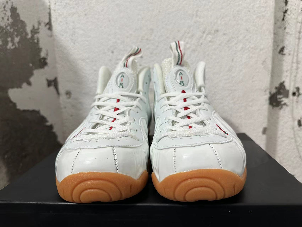 Front view of white Nike Foamposite sneakers with white laces, tongue badges, and gum-colored toe outsoles