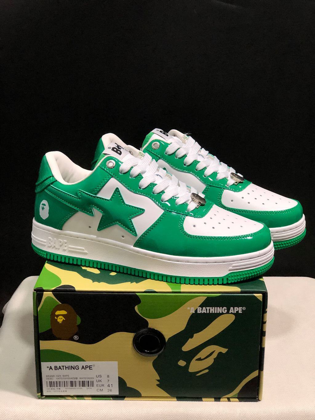 Pair of white and green BAPE STA low-top sneakers displayed on a camouflage-patterned box; the box label shows US 8, UK 7, EU 41, and CM 26 for the photographed sample.