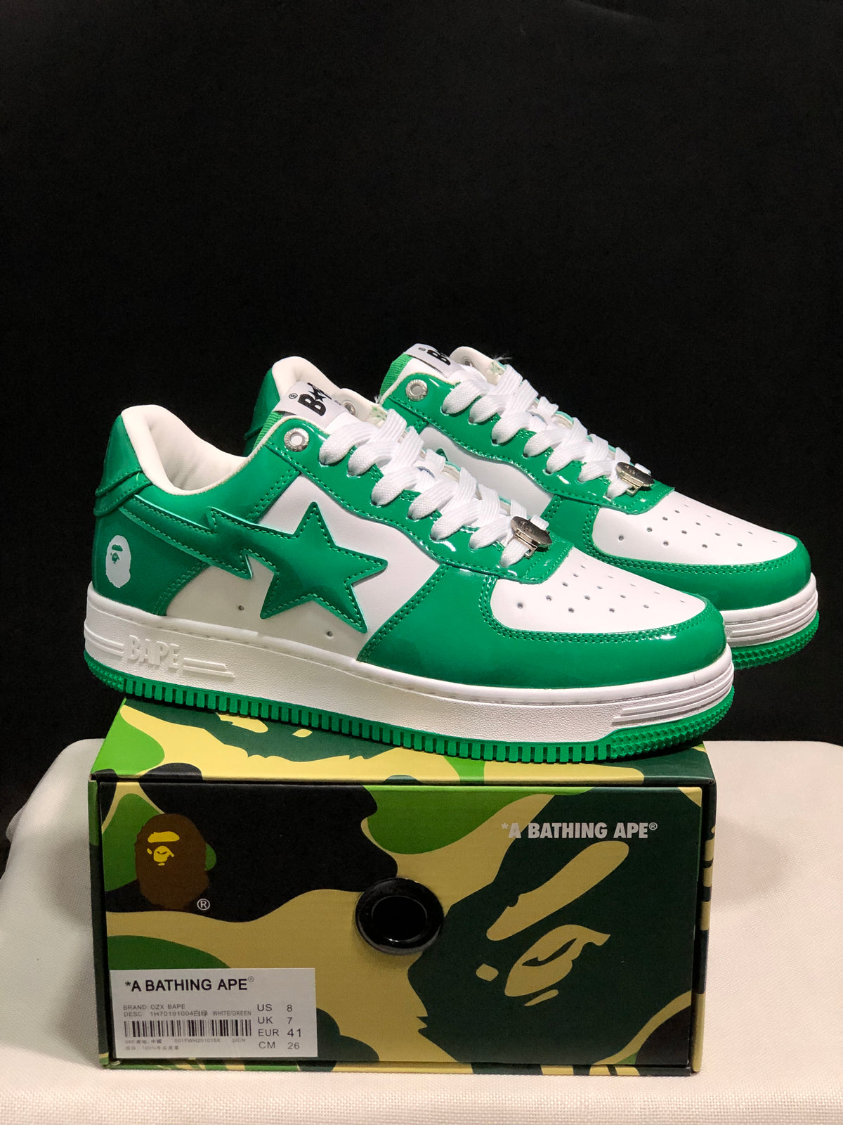 Pair of white and green BAPE STA low-top sneakers displayed on a camouflage-patterned box; the box label shows US 8, UK 7, EU 41, and CM 26 for the photographed sample.
