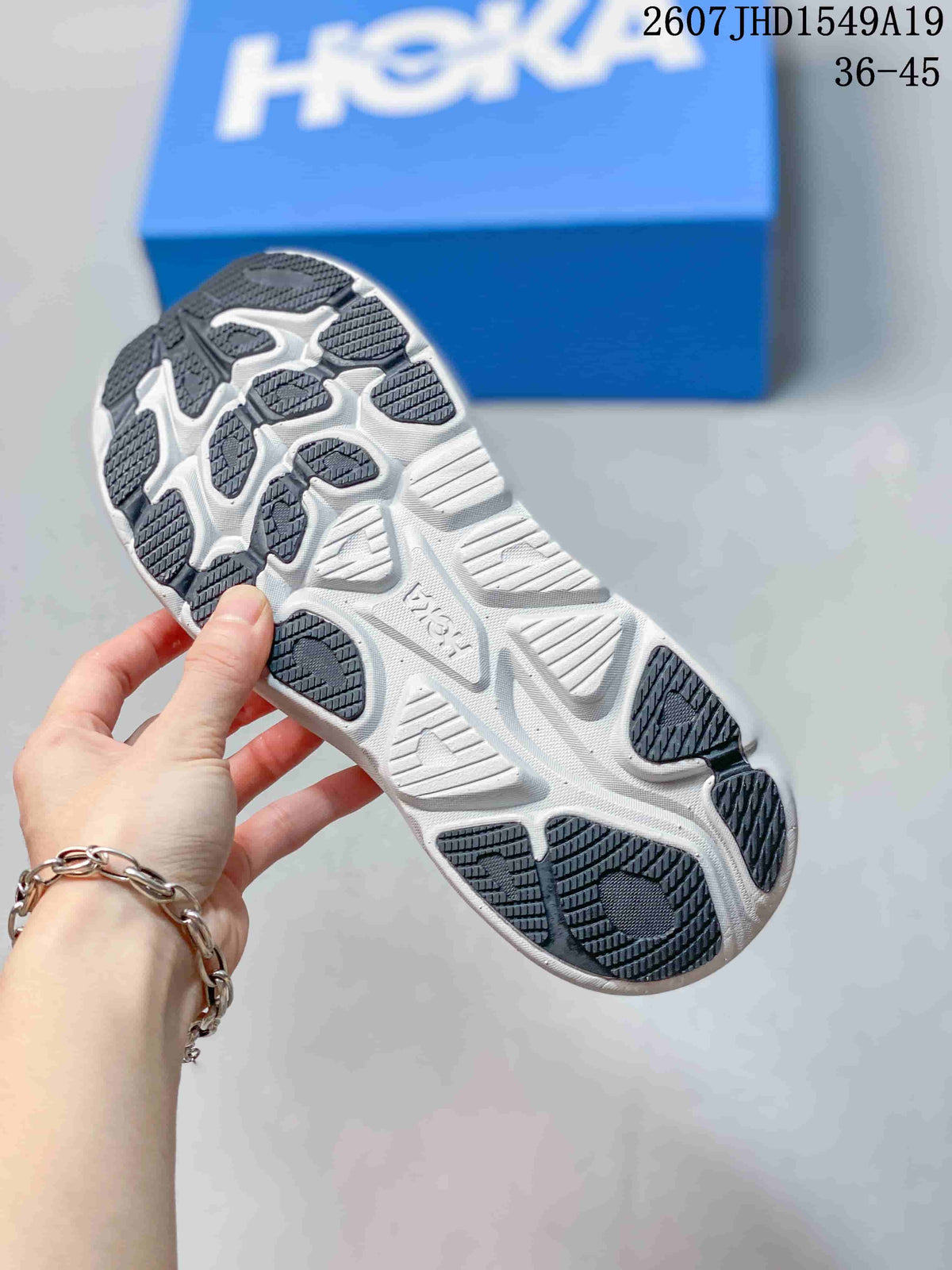 Close view of the sneaker outsole showing a white sole with dark patterned traction sections; a blue Hoka box is in the background.