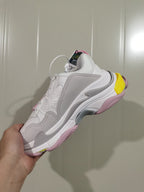 Side view of white Balenciaga Triple S sneakers with grey panels, pink outsole details and a yellow heel accent.