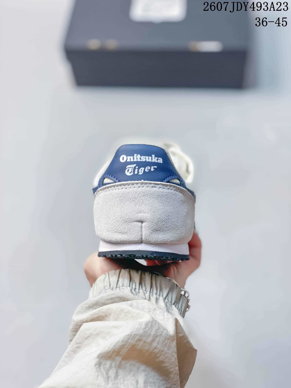 Rear view of a white sneaker showing the padded heel and blue Onitsuka Tiger tongue branding; overlay shows model code 2607JDY493A23 and size range 36–45.