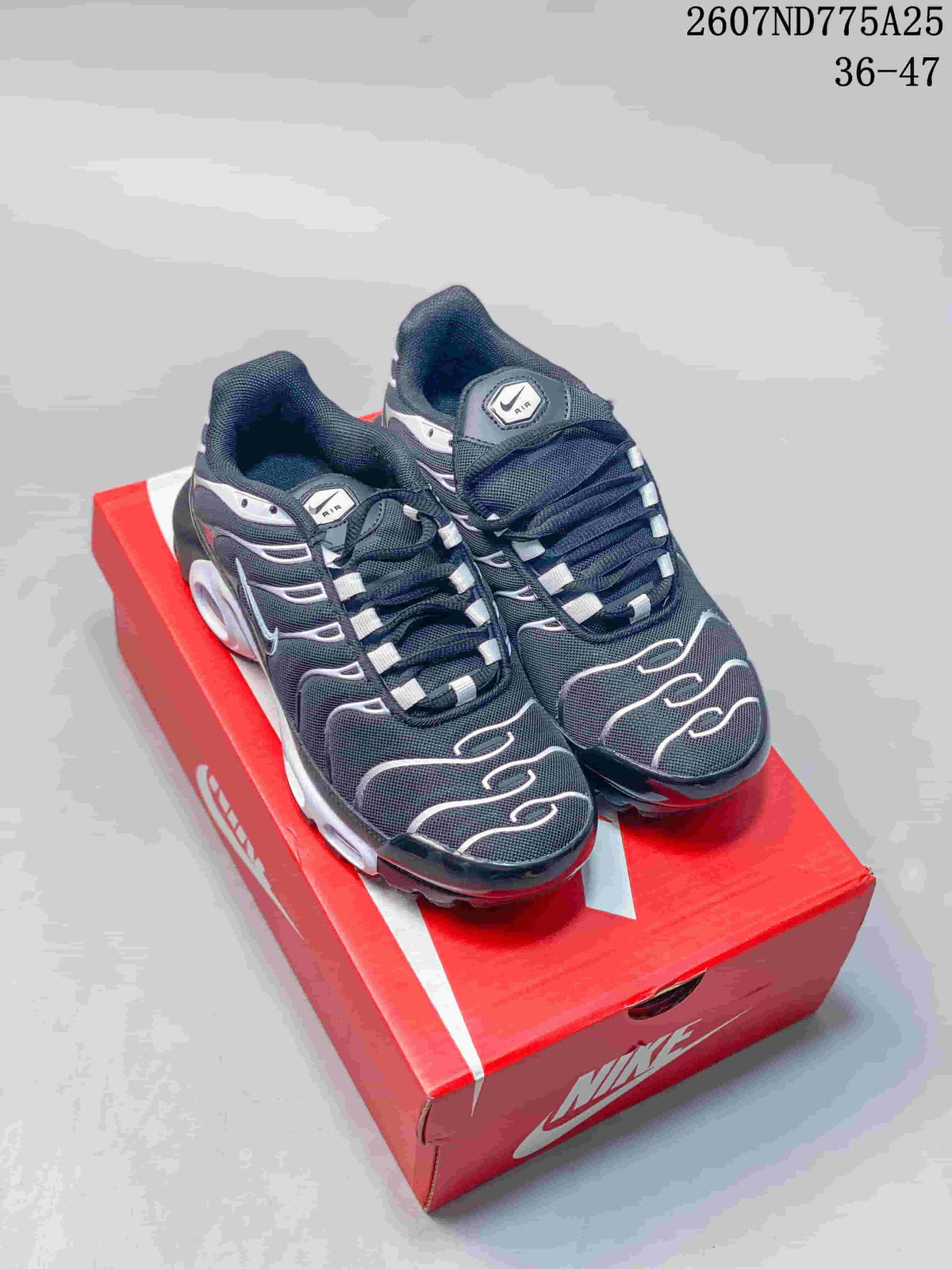 Pair of black and white Nike Air Max Plus TN sneakers displayed on a red Nike shoebox; image text shows 2607ND775A25 and size range 36–47.
