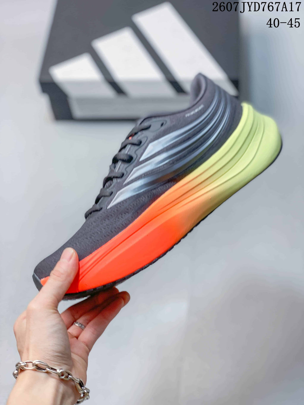 Side profile of an Adidas Supernova Rise sneaker showing the dark gray upper, layered silver detailing, orange midsole edge, and lime-green heel section; image text shows size range 40–45.