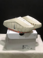 Light bone Nike Mind 001 mules displayed on a shoebox, showing the perforated upper, grey Swoosh, and orange outsole pods.