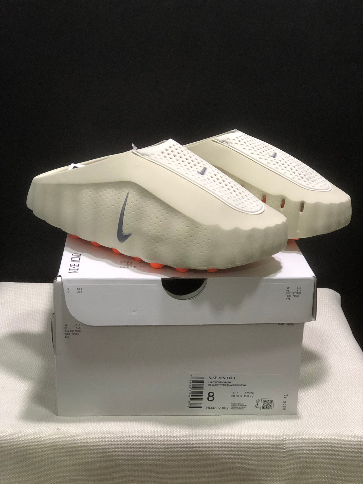 Light bone Nike Mind 001 mules displayed on a shoebox, showing the perforated upper, grey Swoosh, and orange outsole pods.