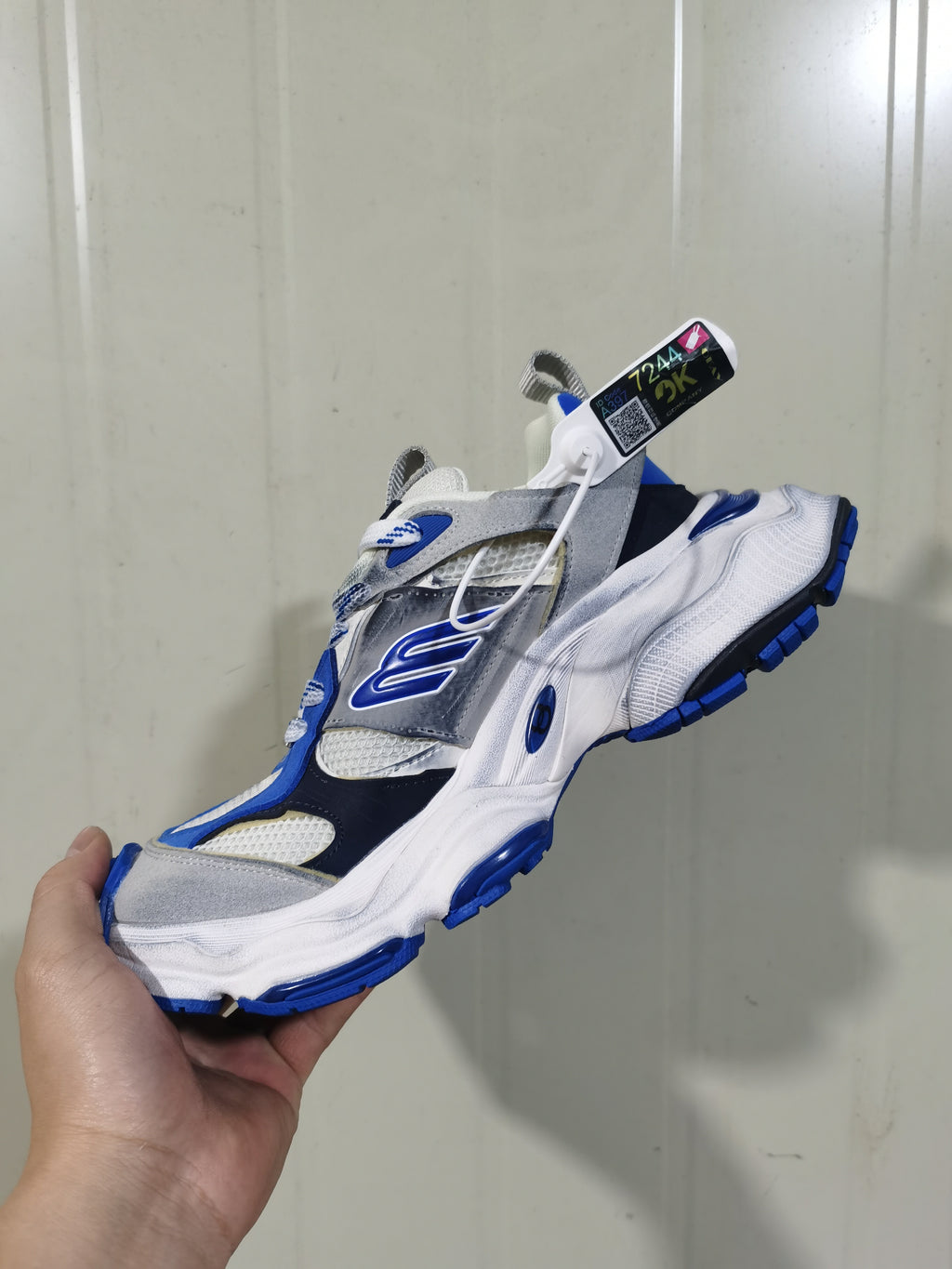 Angled view of the white, grey, and blue sneaker showing its layered upper, side logo, laces, and blue outsole accents.