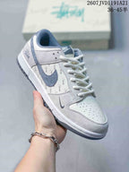 Angled side view showing the white and gray upper, blue-gray Swoosh, repeating Stussy script and dark gray outsole.