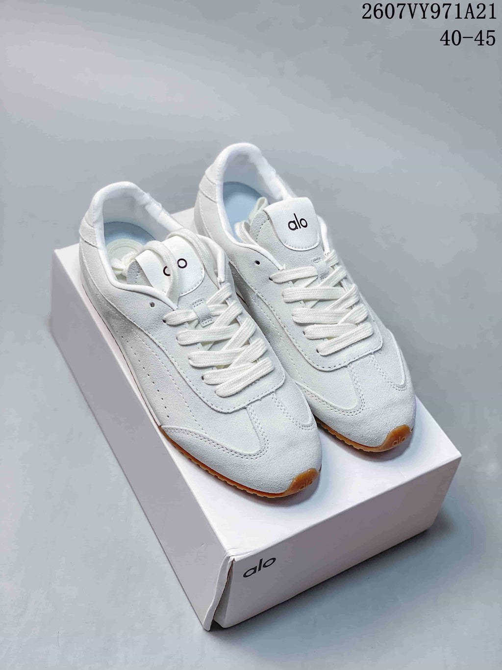 White Alo Yoga low-top sneakers with white laces and gum-colored outsoles displayed on a box; source text shows style code 2607VY971A21 and sizes 40–45.