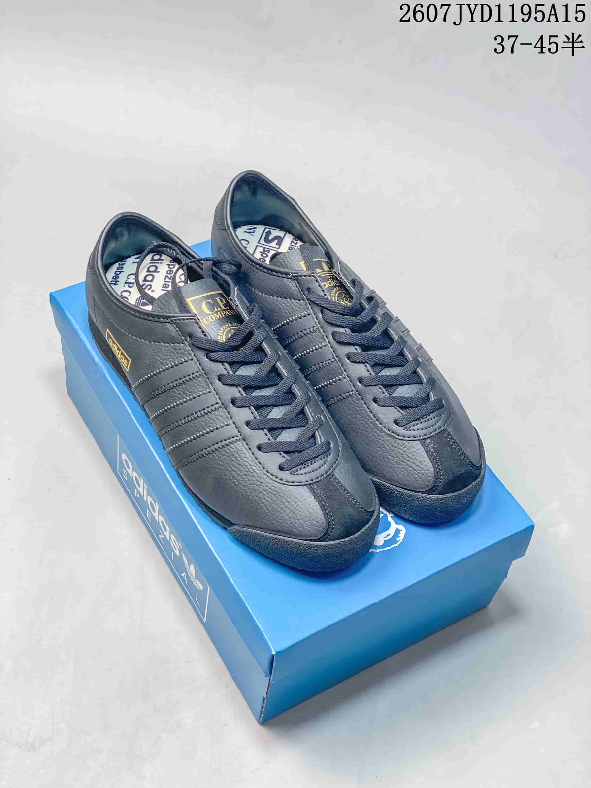 Pair of dark gray adidas C.P. Company sneakers displayed on a blue shoebox, showing the lace-up fronts, tonal side stripes, and gold tongue branding.