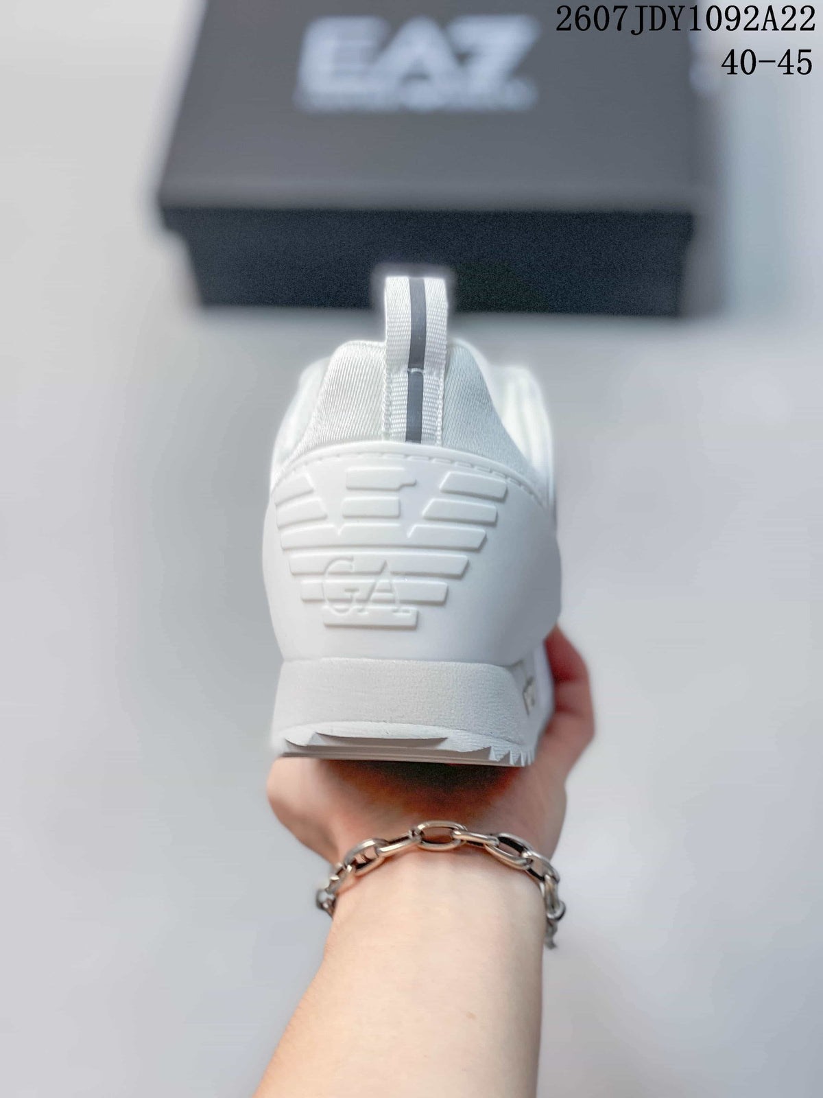 Rear view of a white EA7 sneaker showing the heel pull tab, embossed EA7 mark, and outsole edge; source overlay shows style code 2607JDY1092A22 and size range 40–45.