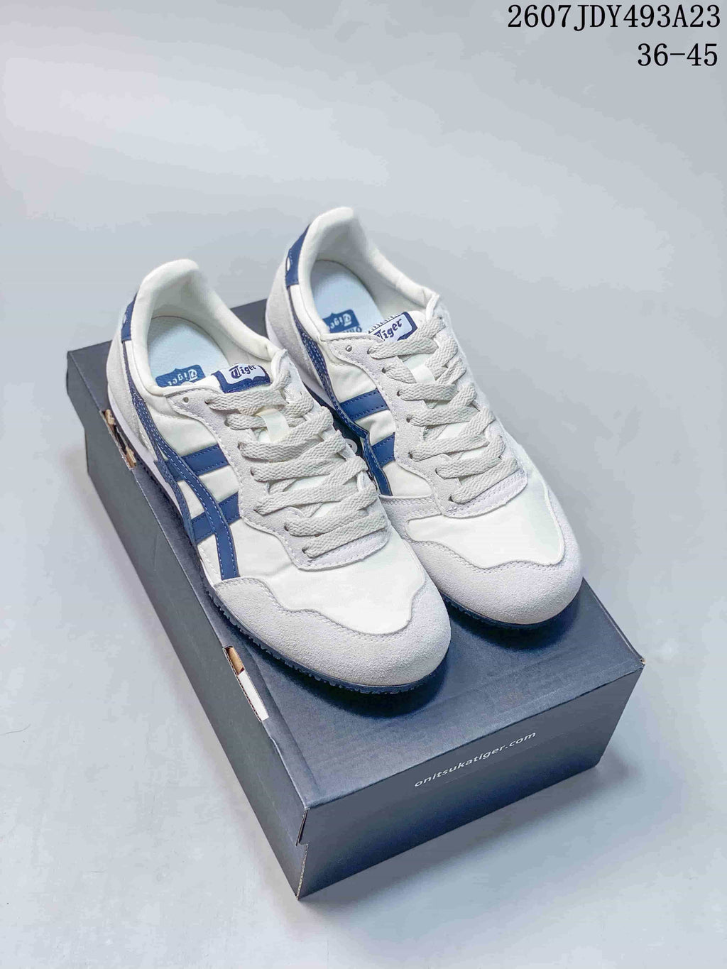 Pair of white and blue Onitsuka Tiger Serrano sneakers displayed on a shoe box; overlay shows model code 2607JDY493A23 and size range 36–45.