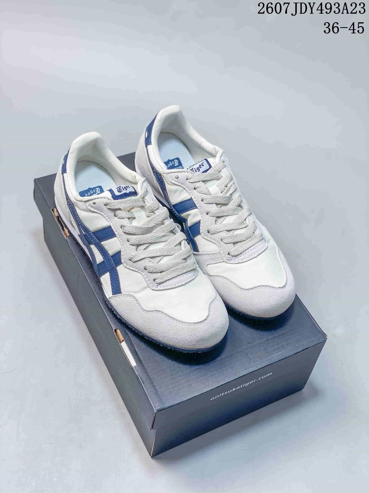 Pair of white and blue Onitsuka Tiger Serrano sneakers displayed on a shoe box; overlay shows model code 2607JDY493A23 and size range 36–45.