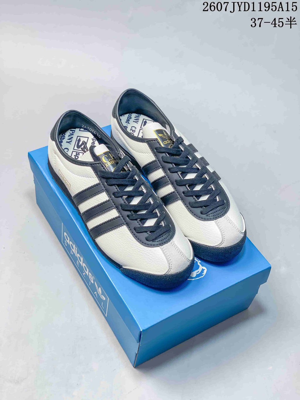 Pair of off-white and navy adidas x C.P. Company sneakers displayed on a blue shoebox.