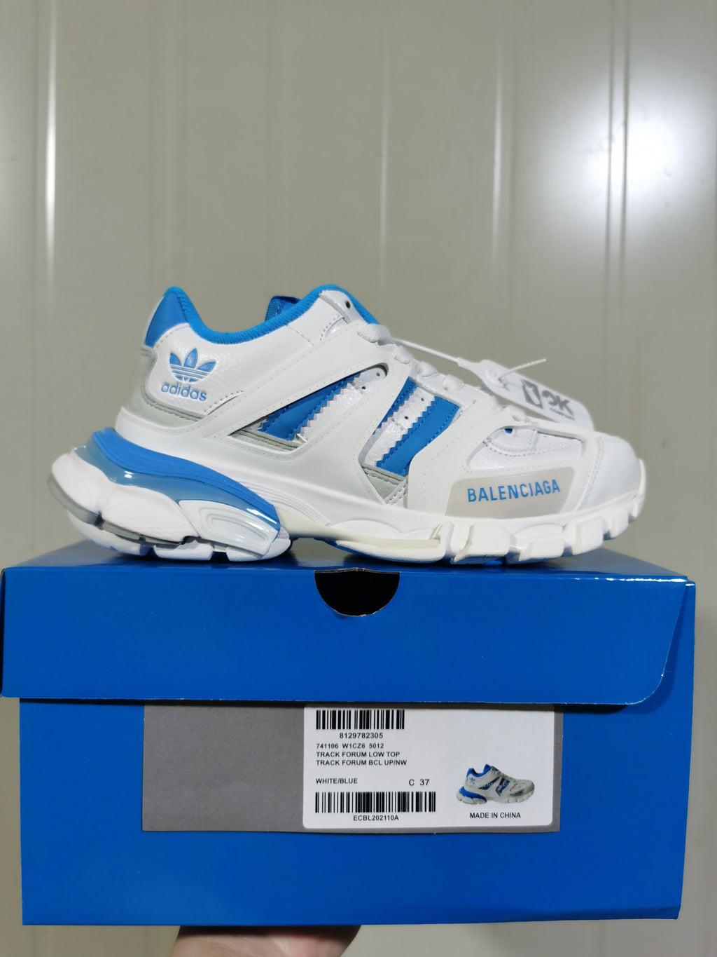 White and blue low-top sneaker displayed on a blue shoe box, with the box label showing Track Forum Low Top and sample size C 37.