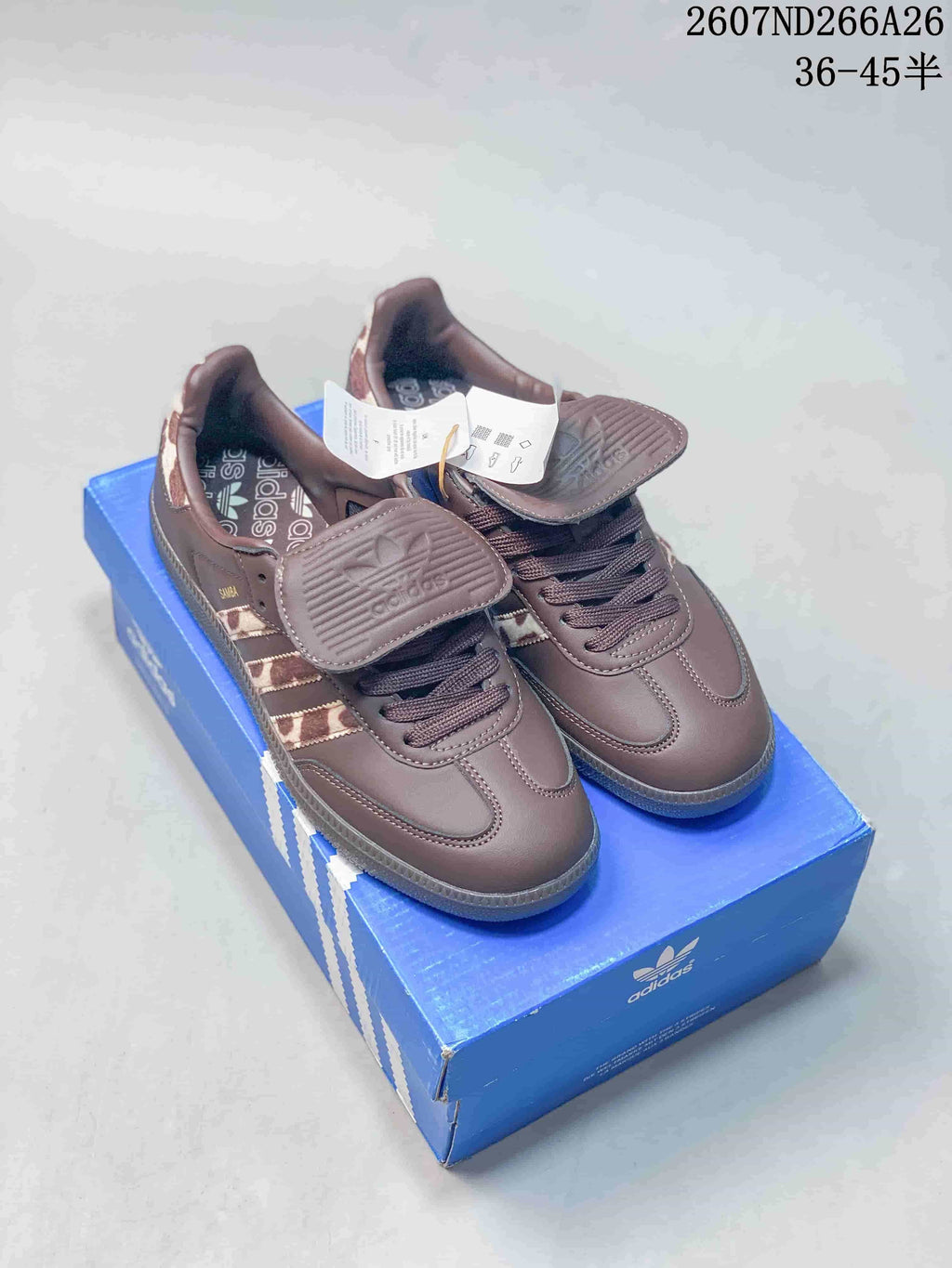 Brown adidas Originals Samba OG sneakers displayed on a blue adidas shoebox, showing the flap tongues, laces, patterned side panels, and branded insoles.