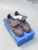 Brown adidas Originals Samba OG sneakers displayed on a blue adidas shoebox, showing the flap tongues, laces, patterned side panels, and branded insoles.