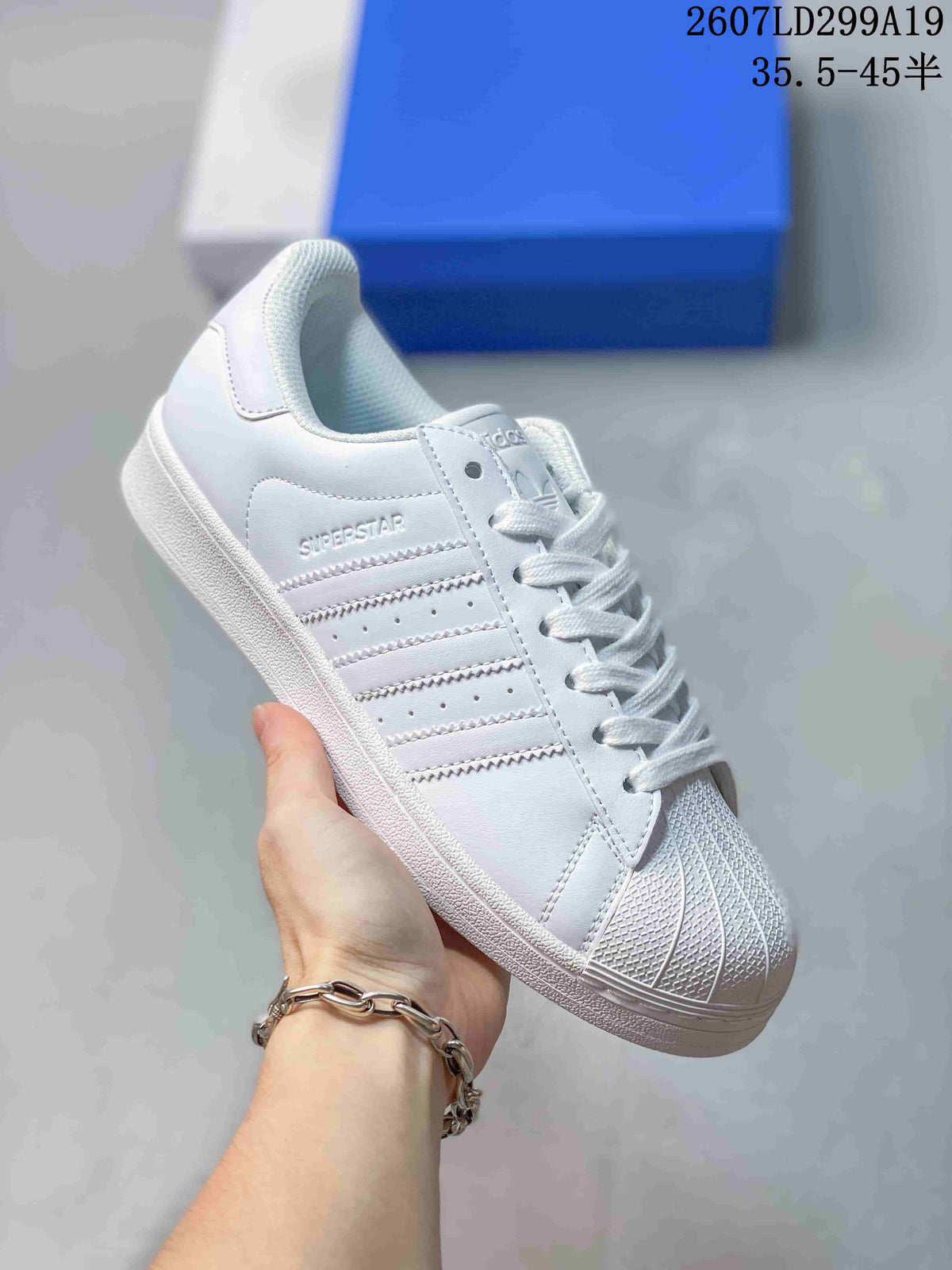 Angled side view of a white Adidas Superstar sneaker showing the shell toe, three stripes and visible Superstar text.