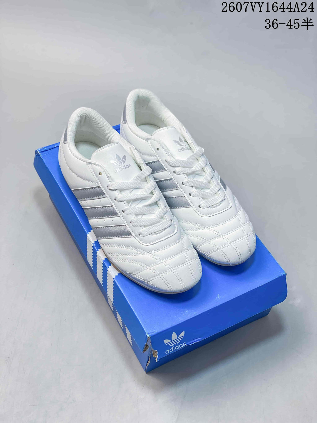 Pair of white and silver adidas Taekwondo sneakers displayed on a blue shoebox.
