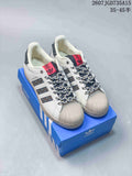Pair of white and black adidas Superstar sneakers displayed on a blue shoebox, showing the shell toes, patterned side stripes, braided laces, and red tongue labels.
