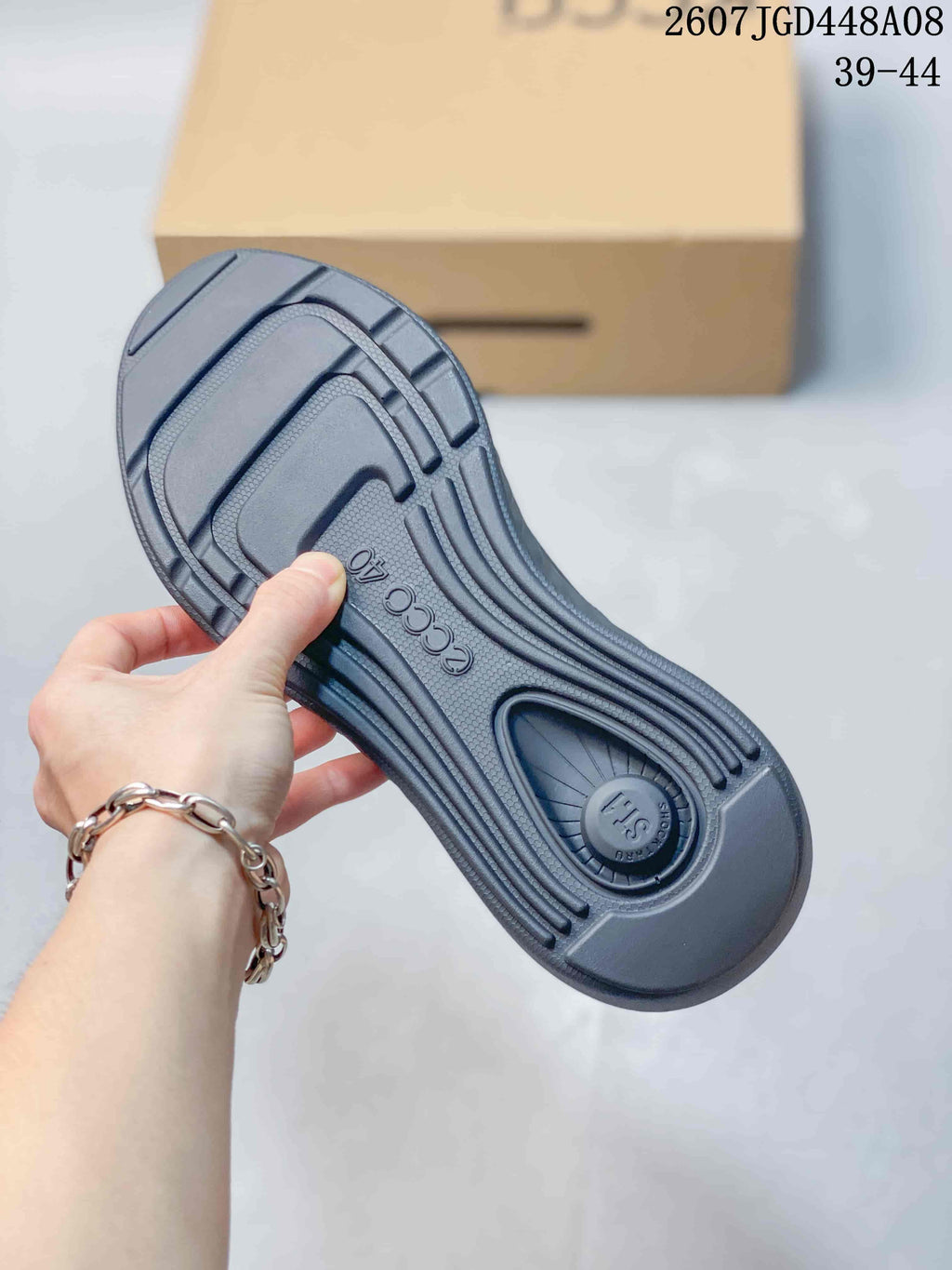 Underside of a dark gray sneaker showing the molded outsole and patterned tread, with product code 2607JGD448A08 and size range 39–44 visible in the background.