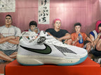 Side profile of a white Nike basketball sneaker with a dark green and black swoosh, patterned side detail, speckled midsole, and blue-green outsole.