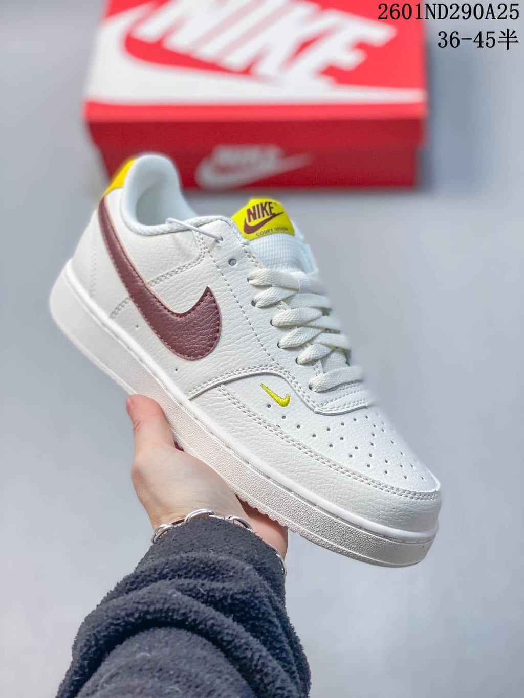 White Nike Court Vision Low sneaker with a burgundy Swoosh and small yellow tongue detail.