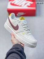 White Nike Court Vision Low sneaker with a burgundy Swoosh and small yellow tongue detail.