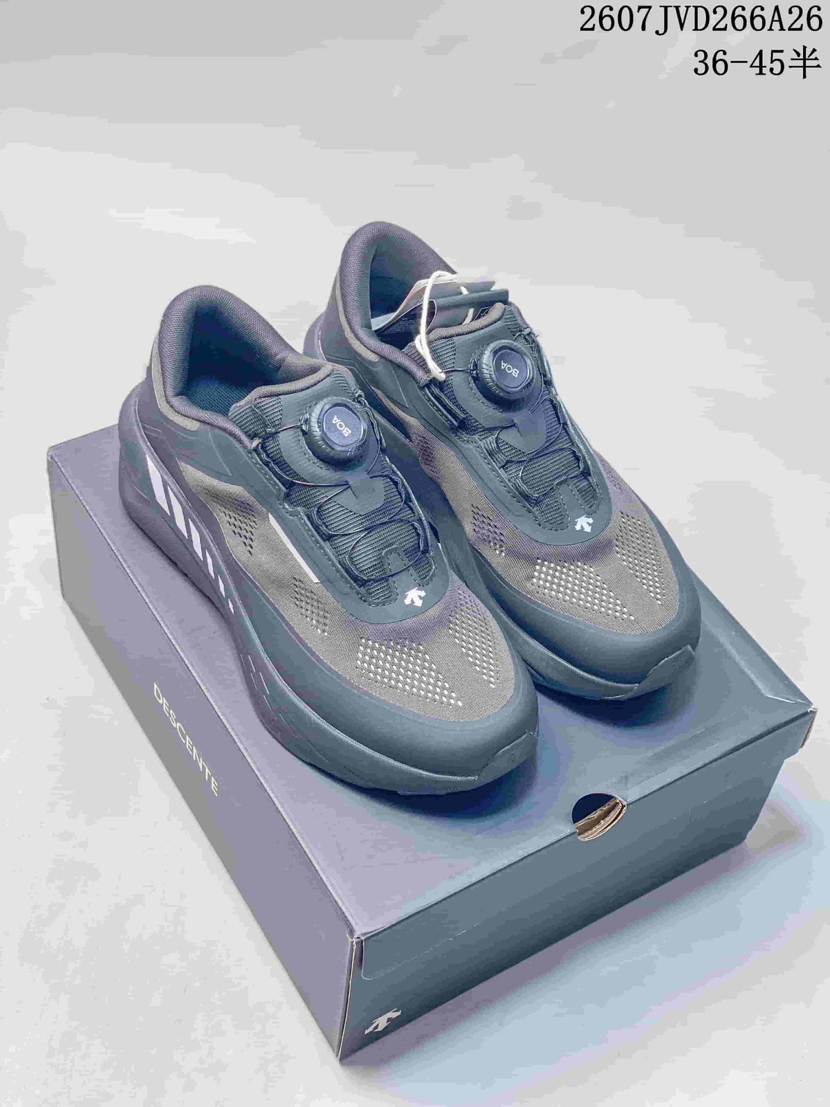 Pair of dark gray Descente Traxis sneakers displayed on a branded shoebox, showing the dial closures and perforated uppers.