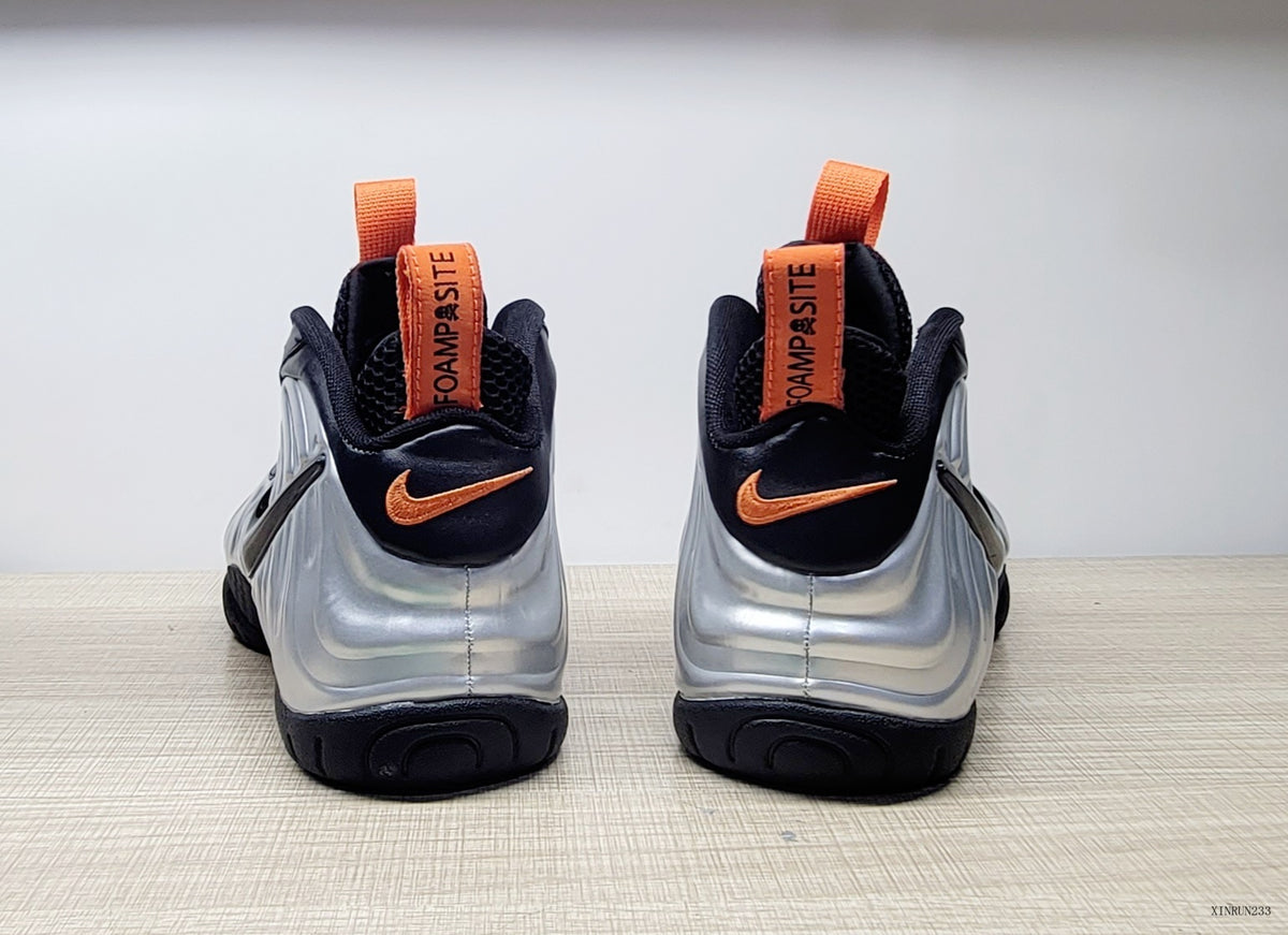 Rear view of silver and black Nike Air Foamposite Pro sneakers with orange pull tabs and accents