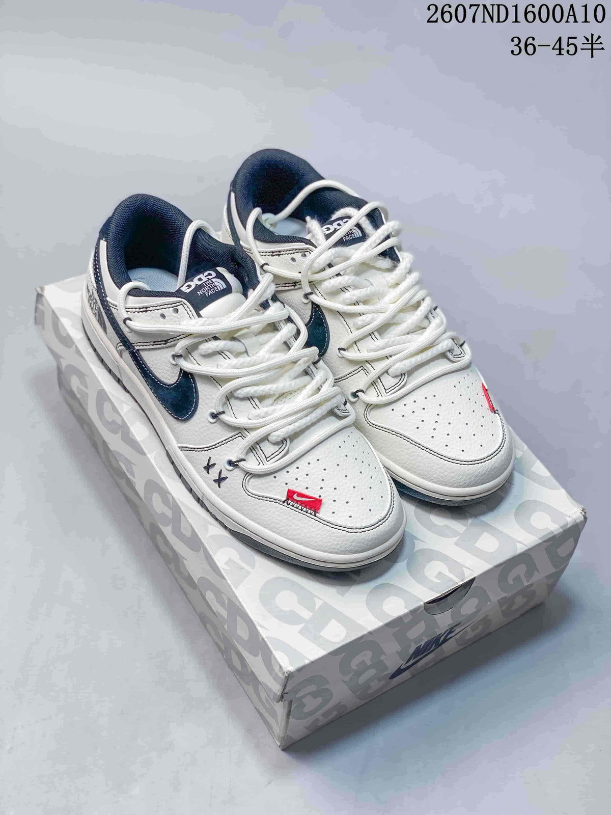 Pair of white and navy Nike SB Dunk Low sneakers displayed on a patterned shoe box, with style code and size range text visible above.